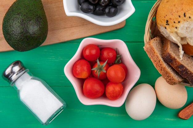 Draufsicht von Tomaten auf einer rosa Schüssel mit Avocado und schwarzen Oliven auf einem hölzernen Küchenbrett mit einem Eimer Brot auf grüner Oberfläche