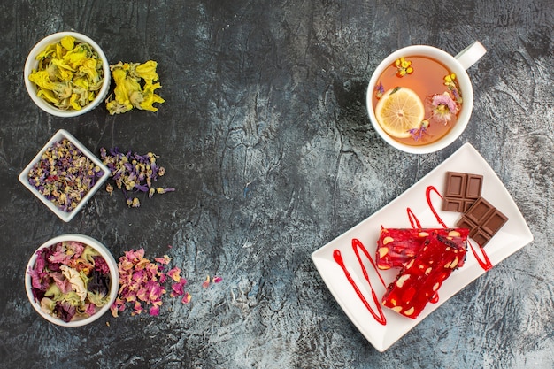 Draufsicht von trockenen Blumen auf Schalen und einer Tasse Kräutertee nahe einem Schokoladenteller auf grauem Grund