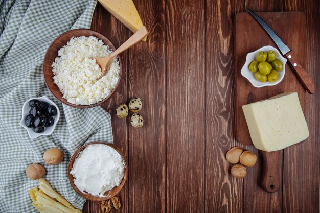 Draufsicht von verschiedenen Käsesorten und Hüttenkäse in einer Schüssel mit Walnüssen, Wachteleiern und eingelegten Oliven auf Holzschneidebrett mit einem Messer auf rustikalem Tisch