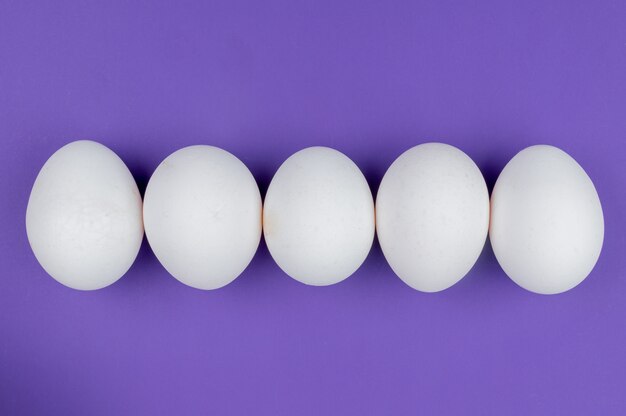 Draufsicht von weißen frischen und gesunden Hühnereiern, die in einer Linie auf einem violetten Hintergrund angeordnet sind