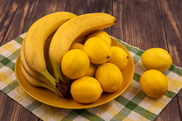 Draufsicht von Zitronen mit abgerundeter Form auf einem gelben Teller auf einem karierten Tuch mit Bananen auf einer Holzoberfläche