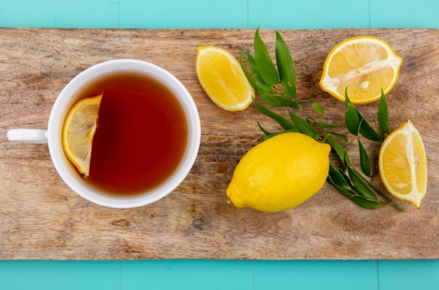 Draufsicht von Zitronen mit Estragon auf einem hölzernen Küchenbrett mit einer Tasse Tee auf blauer Oberfläche