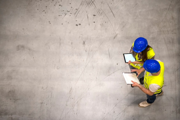 Draufsicht von zwei Industriearbeitern, die Schutzhelme und reflektierende Jacken tragen, die Tablette und Checkliste auf grauem Betonboden halten