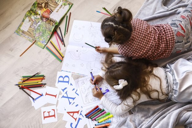 Draufsicht von zwei winzigen Mädchen, die in dem Malbuch zeichnen, das auf dem Boden auf der Decke liegt
