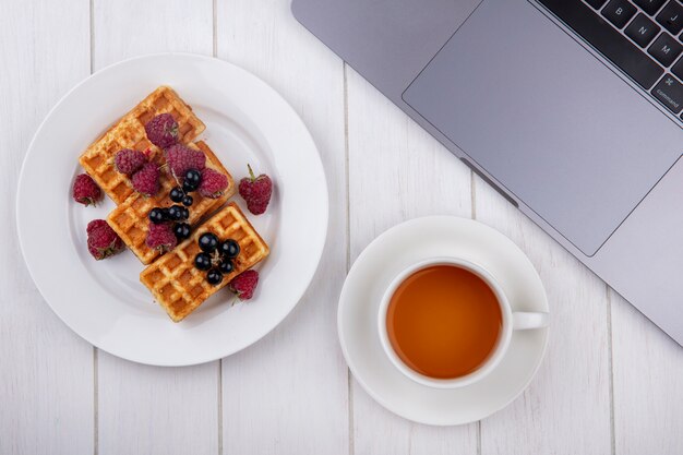 Draufsicht Waffeln auf einem Teller mit Himbeeren und einer Tasse Tee mit einem Laptop auf einem weißen Tisch