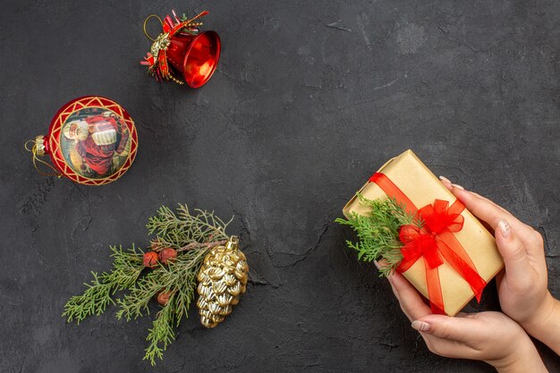 Draufsicht weibliche Hände, die Weihnachtsgeschenk in braunem Papier halten, das mit rotem Band-Weihnachtsbaumschmuck auf dunkler Oberfläche gebunden ist