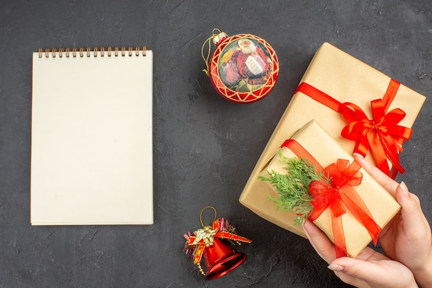 Draufsicht weibliche Hände halten Weihnachtsgeschenk in braunem Papier gebunden mit rotem Band Weihnachtsbaum Spielzeug Notizblock auf dunklem Hintergrund