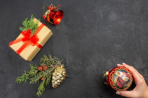 Draufsicht Weihnachtsgeschenk in braunem Papier gebunden mit rotem Band Weihnachtsbaum Ornamente Weihnachtskugel in weiblicher Hand auf dunkler Oberfläche