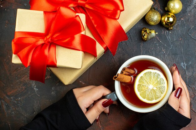 Draufsicht Weihnachtsgeschenke goldene Weihnachtskugeln Tasse Tee in weiblicher Hand auf dunkelrotem Tisch