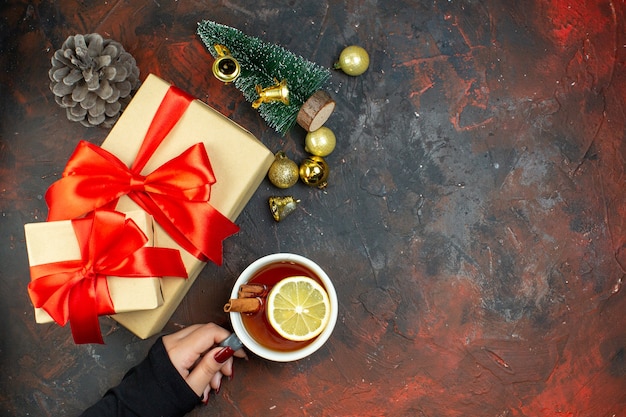 Draufsicht Weihnachtsgeschenke goldene Weihnachtskugeln Tasse Tee in weiblicher Hand Mini-Weihnachtsbaum auf dunkelrotem Tisch freier Platz