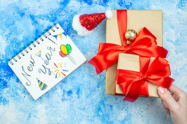 Draufsicht Weihnachtsgeschenke in weiblicher Hand kleine Weihnachtsmütze Neujahr auf Notizblock auf blau-weißem Tisch geschrieben