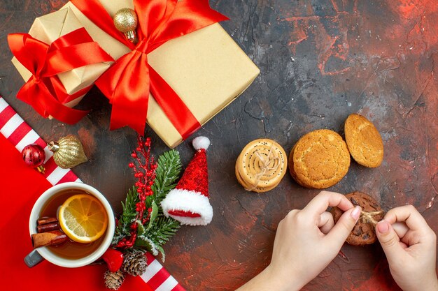 Draufsicht Weihnachtsgeschenke mit rotem Band Weihnachtsmütze Cookies in weiblicher Hand Tasse Tee auf dunkelrotem Tisch gebunden