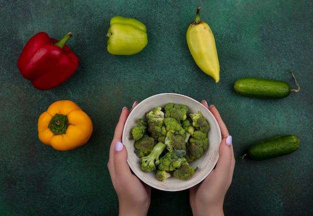 Kostenloses Foto draufsichtfrau, die brokkoli in der schüssel mit gurken und paprika auf grünem hintergrund hält