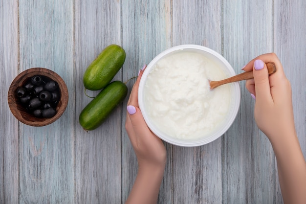 Kostenloses Foto draufsichtfrau, die joghurt in schüssel mit hölzernen löffelgurken und schwarzen oliven auf grauem hintergrund hält