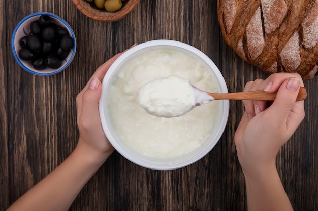 Draufsichtfrau, die Joghurt in Schüssel mit hölzernen Löffeloliven und Schwarzbrot auf hölzernem Hintergrund isst