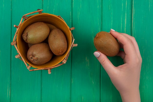 Draufsichtfrau, die Kiwi im Korb auf grünem Hintergrund hält