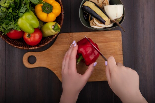 Draufsichtfrau schneidet rote Paprika auf einem Schneidebrett mit Petersilie und Tomaten in einem Korb und Auberginen auf einem hölzernen Hintergrund