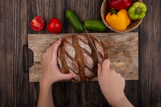 Draufsichtfrau schneidet Schwarzbrot auf einem Ständer mit Tomatengurken und Paprika auf einem hölzernen Hintergrund