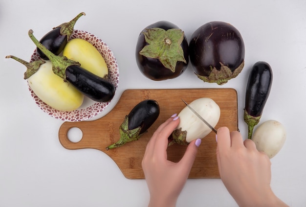 Draufsichtfrau schneidet Schwarzweiss-Aubergine auf einem Schneidebrett mit einem Messer und in einer Platte auf einem weißen Hintergrund