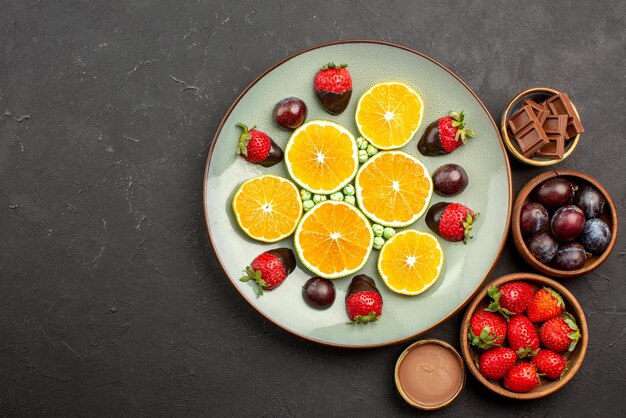 Draufsichtfrüchte auf dem Tisch Erdbeerschokolade und Beeren in Holzschalen neben dem Teller mit gehackten Orangenbonbons und schokoladenüberzogenen Erdbeeren auf dem dunklen Tisch