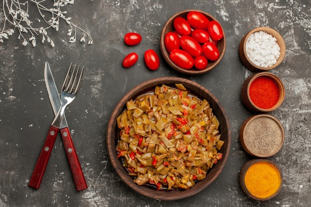 Draufsichtgewürze auf dem Tisch vier Schüsseln mit bunten Gewürzen und Tomaten neben dem Teller mit grünen Bohnen auf dem dunklen Tisch