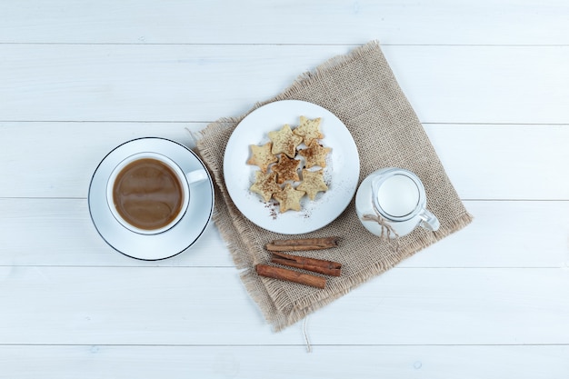Draufsichtkaffee in der Tasse mit Keksen, Zimtstangen, Milch auf Holz und Stück Sackhintergrund.