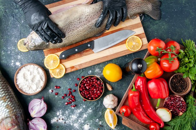 Draufsichtkoch mit schwarzen Handschuhen, die rohen Fisch auf Holzbrett halten Pfeffermühle Mehlschüssel Granatapfelkerne in Schüssel auf dem Tisch