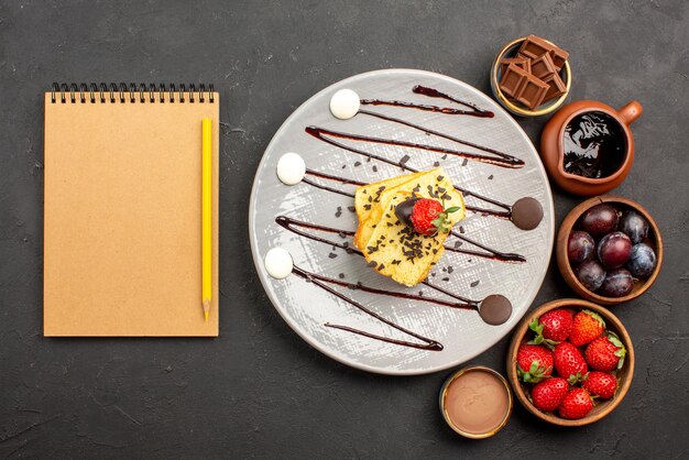 Draufsichtkuchen mit Erdbeer-Erdbeer-Schokolade und Beeren in Schalen und Teller mit Kuchen mit Erdbeeren und Schokoladensauce neben dem Notizbuch und Bleistift auf dem Tisch