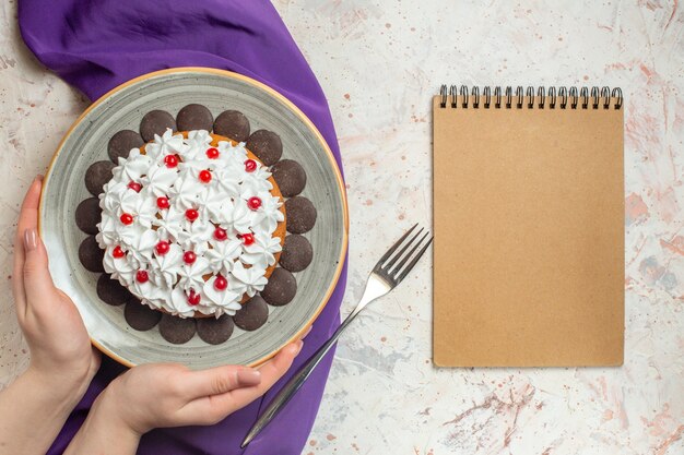 Draufsichtkuchen mit Gebäckcreme auf Teller in der weiblichen Hand lila Schalgabelnotizbuch