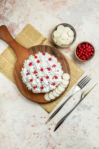 Draufsichtkuchen mit weißer Gebäckcreme auf Holzbrett auf Zeitungsgabel und Tafelmesserbeeren und weißer Schokolade in Schalen