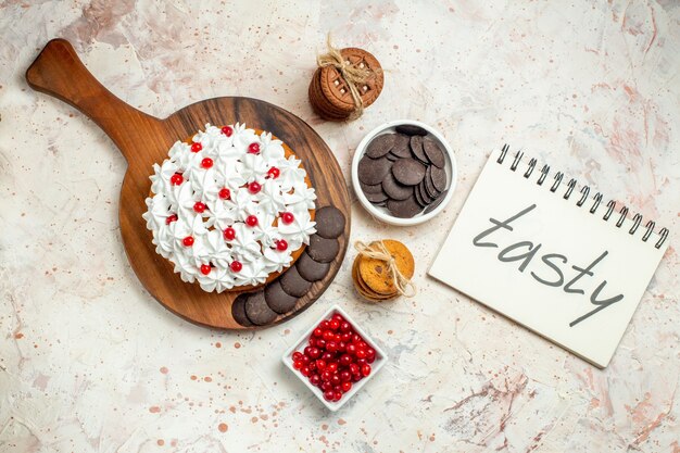 Draufsichtkuchen mit weißer Gebäckcreme auf Schneidebrettschalen mit Beeren und Schokoladenkeksen, die mit einem Seil gebunden sind, lecker auf Notizbuch auf hellgrauem Hintergrund geschrieben