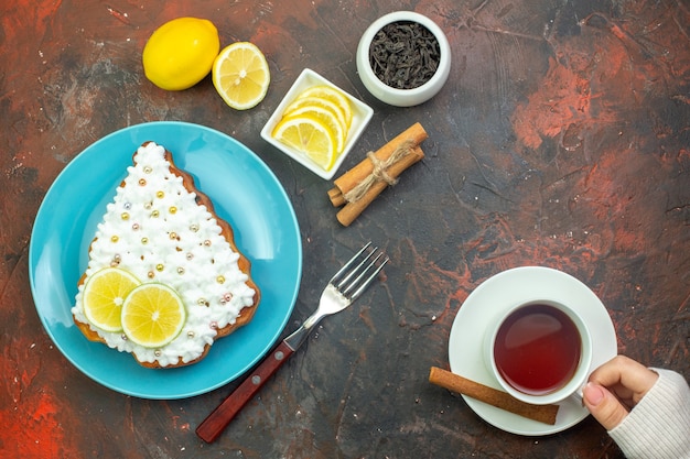 Draufsichtkuchen mit Zitrone auf blauem Teller Zitronenscheiben in Schüsselgabel Tasse Tee in weiblicher Hand Zimtstangen auf dunkelrotem Hintergrund