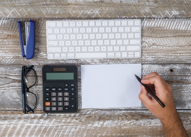 Draufsichtmann, der etwas auf Papier schreibt, mit Tastatur, Brille, Stift, Kaktus, Lupe auf hölzernem Hintergrund. horizontal