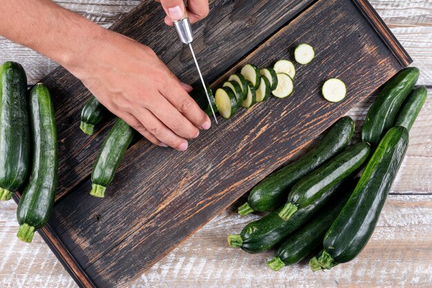 Draufsichtmann, der frische Zucchini in Scheiben auf Schneidebrett auf hellem Holztisch schneidet