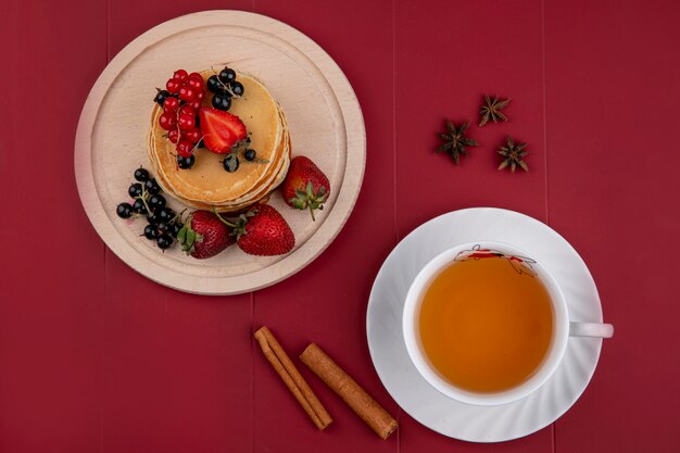 Draufsichtpfannkuchen mit roten und schwarzen Johannisbeeren und Erdbeeren auf einem Tablett mit einer Tasse Tee und Zimt auf einem roten Hintergrund