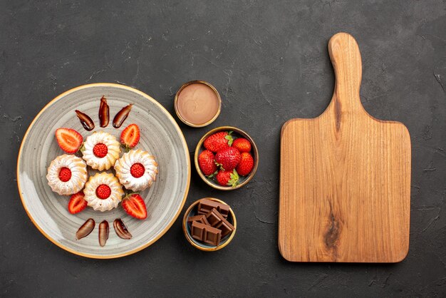 Kostenloses Foto draufsichtplätzchen und erdbeerplätzchen mit erdbeeren auf weißem teller neben schüsseln mit erdbeerschokolade und schokoladencreme neben dem schneidebrett