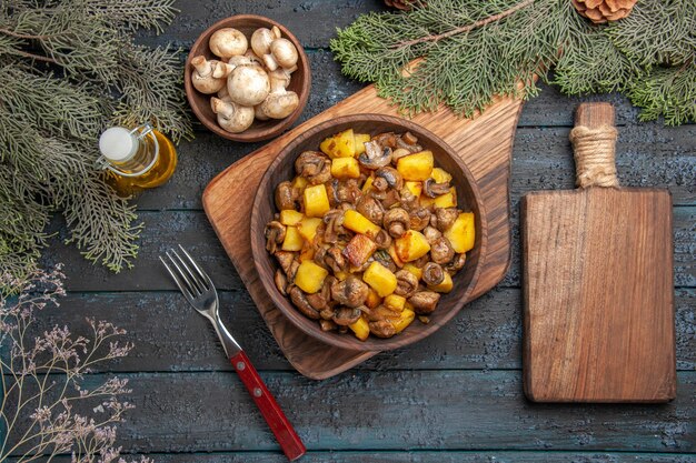 Draufsichtplatte und Schneidebrett Kartoffeln und Pilze in Schüssel auf braunem Brett neben der Gabel und Holzschneidebrett unter Schüssel mit Pilzen Öl in Flasche und Zweigen mit Zapfen