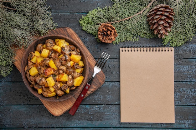 Draufsichtschale und Notizbuch Holzschüssel mit Kartoffeln mit Pilzen auf dem Schneidebrett neben dem Notizbuch und Gabel unter Fichtenzweigen mit Zapfen