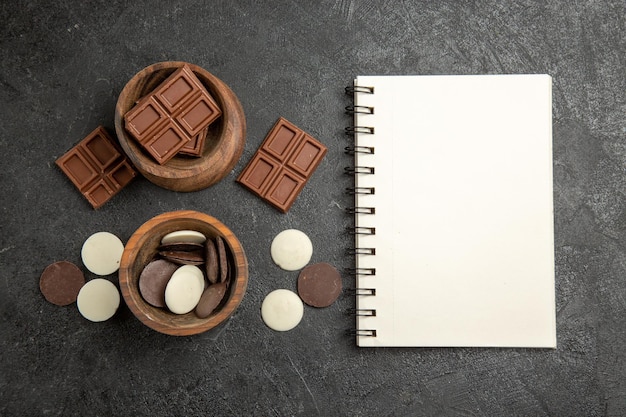 Draufsichtschokolade auf dem Tisch braune Schokoladenschalen neben dem weißen Notizbuch auf dunklem Hintergrund