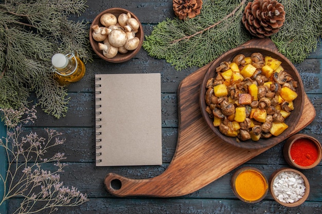 Draufsichtschüssel mit Speiseteller mit Pilzen und Kartoffeln auf dem Schneidebrett neben dem Notizbuch zwischen der Flasche Ölschüssel mit weißen Pilzen, Fichtenzweigen und bunten Gewürzen