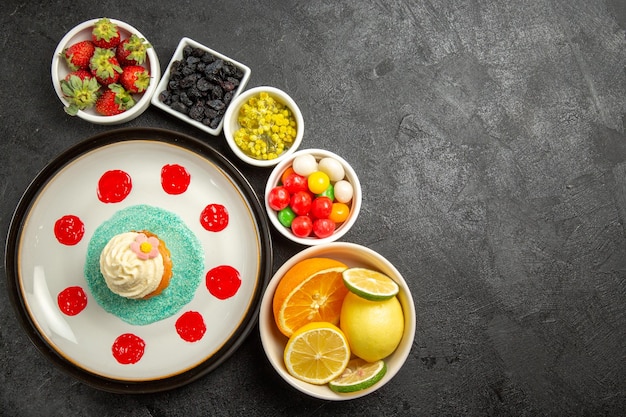 Draufsichtschüsseln mit Süßigkeiten Schüsseln mit verschiedenen Süßigkeiten, Zitrusfrüchten und Beeren neben dem Kuchenteller mit Sahne und Soßen auf dem schwarzen Tisch