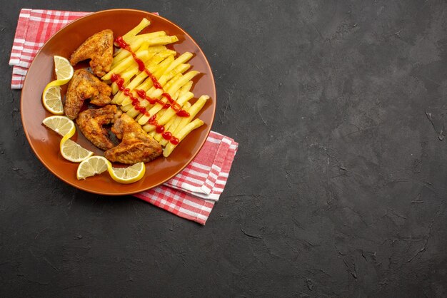 Draufsichtteller auf Tischdecke oranger Teller mit appetitlichen Pommes Frites Chicken Wings Ketchup und Zitrone auf rosa-weiß karierter Tischdecke auf der linken Seite des dunklen Tisches