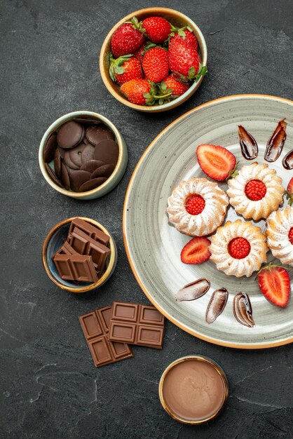 Draufsichtteller mit Keksen Teller mit Erdbeerkeksen mit Schokolade und Schalen mit Schokoladenerdbeere und Schokoladencreme auf dunkler Oberfläche