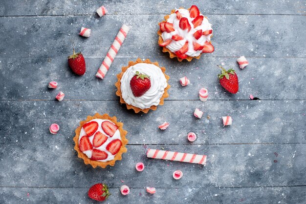 Draußen entfernte Ansicht des kleinen cremigen Kuchens mit geschnittenen und frischen Erdbeeren zusammen mit Stockbonbons auf grauem süßem Auflauf des Fruchtbeerenkuchens