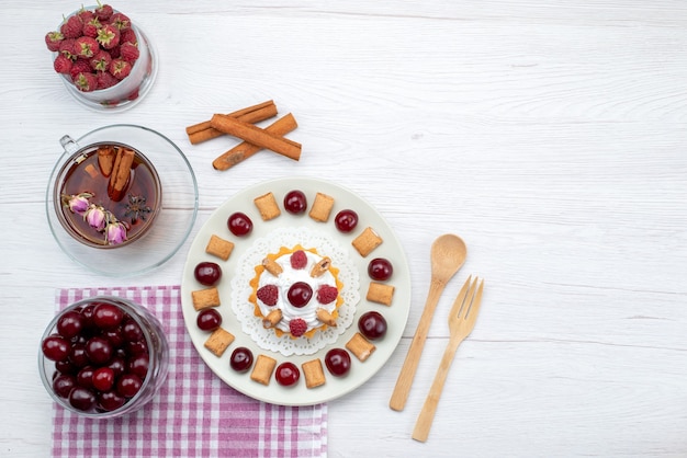 Draußen entfernte Ansicht des kleinen cremigen Kuchens mit Himbeerkirschen und kleinen Kekse-Tee-Zimt auf weißem Schreibtisch, Obstkuchen-Beeren-Sahne-Zucker-Tee