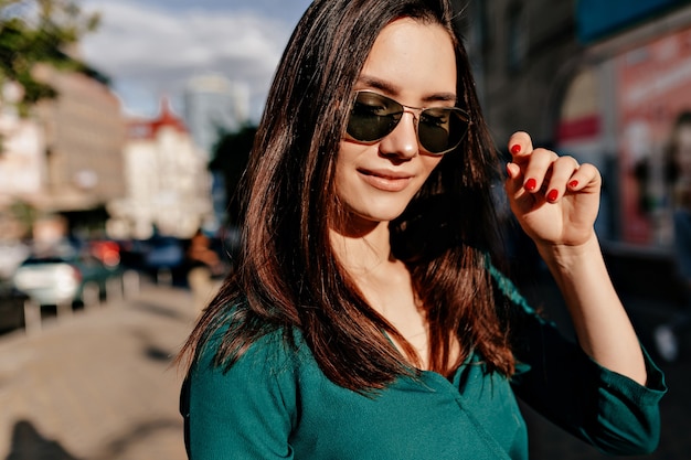 Draußen Nahaufnahmeporträt der bezaubernden europäischen Frau, die schwarze Sonnenbrille und grüne Bluse aufwirfend aufwirft