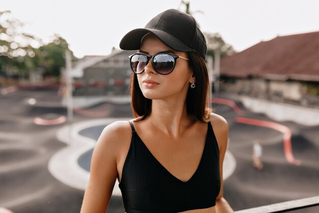 Draußen Nahaufnahmeporträt der stilvollen hübschen Frau in der Sommermütze und in den Gläsern, die während des Fotoshootings draußen im Sonnenlicht auf Skatepark aufwerfen