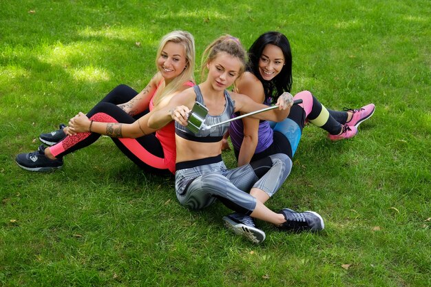 Drei attraktive sportliche Frauen sitzen auf einem Rasen und machen nach dem Fitnesstraining ein Selfie.