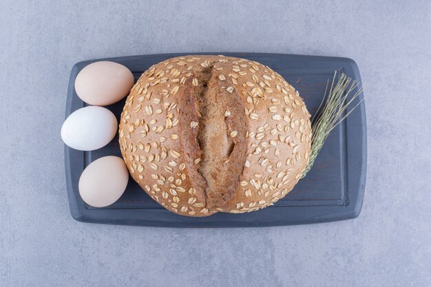 Drei Eier, ein Laib Brot und ein einzelner Weizenstiel auf einem Brett auf Marmoroberfläche