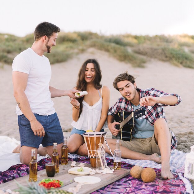 Drei Freunde sitzen am Strand mit Gitarre und Getränken